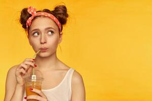 Young lady, pretty ginger woman with two buns and doted hairband. Wearing white shirt and wonder while making a sip of juicy fresh. Watching to the right at copy space over yellow background photo