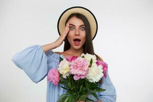 Young lady, pretty woman with long brunette hair. Wearing a hat and blue dress. Holding bouquet of flowers and touching her head, shocked. Watching at the camera isolated over white background photo