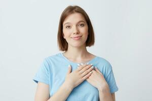 Closeup of smiling charming young woman wears blue t shirt keeps hands on her heart area and feels happy isolated over white background Girl in love photo