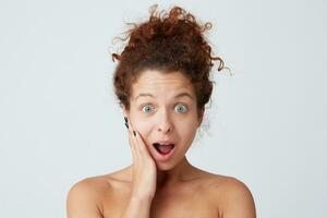 Cropped shot of impressed astonished young woman with curly hair, perfect healthy skin and opened mouth touching face by hand after applying mask or cream and looks amazed isolated over white wall photo