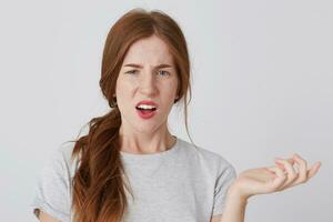 Unhappy confused young woman with red hair and freckles looks displeased and holding copy space on palm isolated over white background photo