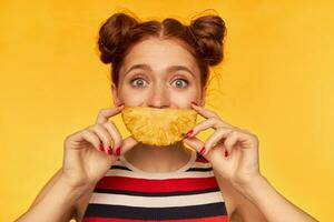 Teenage girl, happy looking red hair woman with two buns and big eyes. Wearing striped tank top and holding slice of a pineapple. Watching at the camera, isolated closeup over yellow background photo