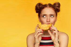 Young lady, pretty ginger woman with two buns and healthy skin. Wearing striped tank top and biting slice of a pineapple. Watching to the left at copy space, isolated closeup over yellow background photo