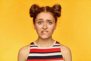 Young lady, pretty ginger woman with two buns. Wearing striped shirt and bite her lip. Looking worried, afraid. Watching at the camera, closeup, isolated over yellow background photo