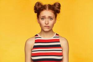 Teenage girl, happy looking red hair woman with two buns. Wearing striped tank top and bite her lip, pending of what you will say. Watching at the camera isolated over yellow background photo