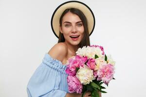 Portrait of attractive, nice looking girl with big smile and long brunette hair. Wearing a hat and blue dress. Holding bouquet of flowers and watching at the camera isolated over white background photo