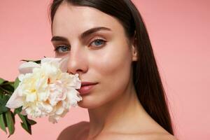 Nice looking woman, beautiful girl with long brunette hair and healthy skin, holding a flower next to her face. Watching at the camera, closeup, isolated over pastel pink background photo