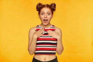 Portrait of attractive, red hair girl with two buns. Wearing striped tank top and looking shocked, put hands on a chest. Emotional concept. Watching at the camera isolated over yellow background photo