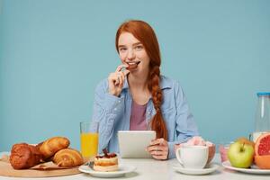 Beautiful smiling red-haired girl having breakfast reding news in her tablet looking camera eating chocolate cookie, isolated on blue background photo