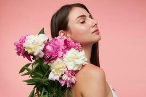 Nice looking woman, beautiful girl with long brunette hair, closed eyes and healthy skin. Wearing white dress and holds bouquet of flowers over here back. Stand isolated over pastel pink background photo