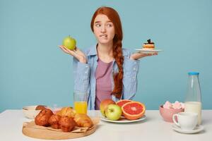 Hard choice. Portrait of beautiful woman trying to choose between healthy and unhealthy food while isolated on bluebackground. Woman holding cake and apple in hands photo