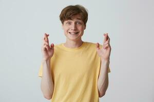 retrato de contento emocionado joven hombre estudiante con metal tirantes en dientes en amarillo t camisa hace un deseo y mantiene dedos cruzado aislado terminado blanco antecedentes cree en milagro foto