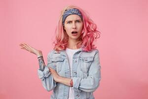 Young beautiful outraged pink haired woman in denim jacket, frown and indignant looking at the camera, wide open mouth in misunderstanding expression. Stands over pink background. photo