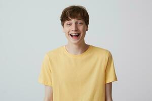 Cheerful blond young man with short haircut and metal braces on teeth wears yellow t shirt and looks happy isolated over white background Looks in camera and laughing photo