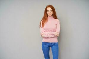 Horizontal photo of beautiful long haired redhead lady dressed in pink knitted sweater and jeans standing over grey background, keeping hands folded on her chest and smiling positively