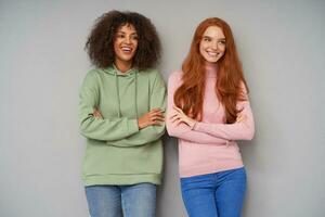 Studio photo of two beautiful young cheerful ladies keeping their hands folded while posing over grey background, looking happily aside and smiling widely
