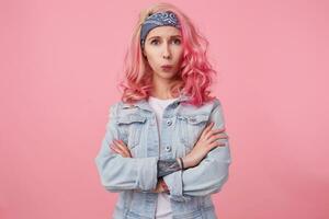 Portrait of young sad beautiful pink haired lady in denim jacket with crossed arms, displeased looks at the camera, someone told her something offensive, stands over pink background. photo