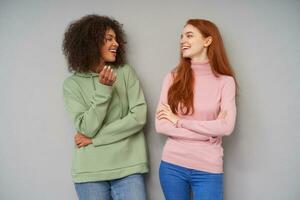 Shot of two joyful attractive young ladies with casual hairstyle smiling cheerfully while having pleasant conversation, standing against grey background and looking on each other photo