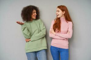 Indoor photo of pretty positive young girlfriends looking cheerfully on each other with pleasant smile while sharing news, wearing casual clothes while posing over grey background