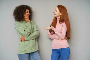 Cheerful young lovely ladies smiling positively while meeting each other and having pleasant talk, wearing comfortable casual clothes while posing over grey background photo