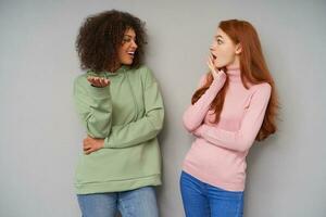 Horizontal photo of young attractive women having exciting conversation while posing over grey background, raising emotionally their hands while sharing news