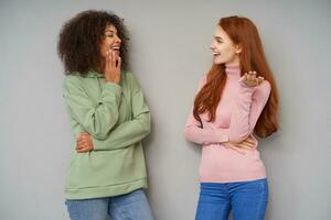 Indoor shot of pretty happy ladies dressed in casual clothes looking on each other while having pleasant talk and smiling cheerfully, isolated over grey background photo