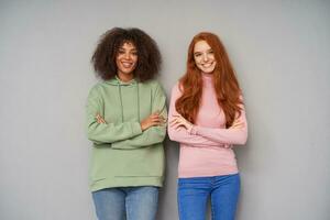 Two attractive young cheerful ladies dressed in casual clothes looking positively at camera and smiling widely, crossing their hands on chest while posing over grey background photo