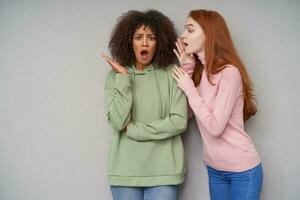 Shocked young dark haired curly female with dark skin looking at camera with wide mouth opened and raising amazedly palm while hearing unexpected news, isolated over grey background photo