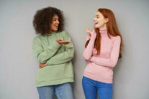 Young beautiful joyful ladies with casual hairstyle keeping hands raised while talking to each other and smiling cheerfully, standing against grey background in cosy clothes photo