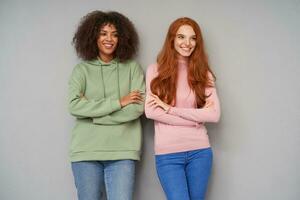 Pair of young happy pretty women in casual wear being in nice mood and smiling broadly while standing over grey background, crossing their hands on chest photo