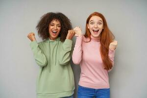 Portrait of joyful young lovely ladies looking excitedly at camera and keeping mouths wide opened, clenching raised hands and screaming happily while posing over grey background photo