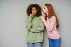 Agitated young brown-eyed curly dark skinned brunette woman covering her mouth and frowning eyebrows while her girlfriend telling her exciting news, standing against grey background photo