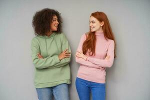 Studio photo of charming cheerful young ladies looking positively on each other and smiling happily, keeping hands crossed on chest while posing over grey background