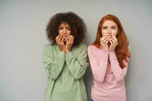 Frightened young attractive women looking scaredly to camera and biting undeplis while standing over grey background, holding their faces with raised hands photo