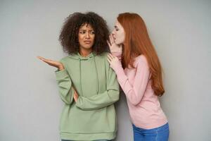 Confused young pretty curly dark skinned brunette female raising palm and frowning her puzzled face while hearing story from her long haired redhead girlfriend, isolated over grey background photo