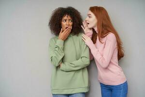 Indoor photo of confused young dark haired curly female with dark skin raising palm to her mouth while listening to shocking news from her redhead long haired lovely girlfriend