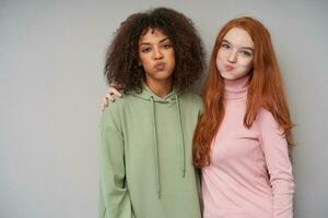 Funny shot of lovely cheerful ladies looking positively at camera while puffing out their cheeks and embracing each other, standing against grey background photo