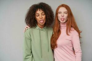 Portrait of cheerful attractive women keeping their eyes closed while fooling and showing tongue at camera, hugging each other while standing over grey background photo