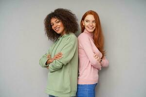 Joyous attractive young women showing their pleasant emotions and smiling cheerfully while standing over grey background with crossed hands, dressed in casual wear photo