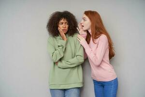 Puzzled young curly dark skinned brunette lady in green hoodie and jeans keeping palm on her cheek and frowning confusedly eyebrows while hearing unexpected story from her friend photo