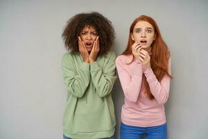 Afraid young pretty girlfriends raising hands to their shocked faces while looking scaredly at camera, posing over grey background in comfortable casual wear photo