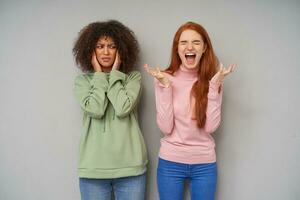 Stressed young attractive ladies in casual clothes frowning their faces and expressing negative emotions while standing posing over grey background, having bad day photo