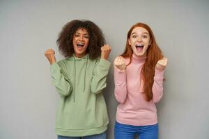 Overjoyed young lovely attractive women raising happily their fists while looking emotionally at camera with wide mouths opened, isolated over grey background photo