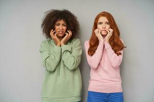 Shocked young pretty ladies dressed in casual clothes keeping raised hands on their cheeks while looking scaredly at camera, standing against grey background photo