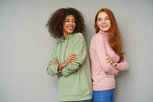 Indoor photo of lovely cheerful young ladies showing their white perfect teeth while smiling positively, folding hands on their chests while posing over grey background