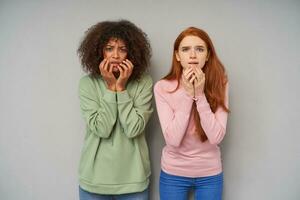 Scared young beautiful ladies in casual clothes frowning their eyebrows and keeping raised hands on their frightened faces while standing over grey background photo