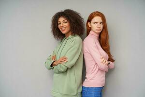 Horizontal shot of beautiful young girlfriends with casual hairstyle expressing different emotions while standing over grey background with crossed hands photo