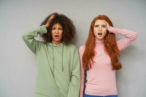 Confused young pretty women holding raised hand on their heads while standing over grey background, frowning eyebrows and keeping mouths opened while looking at camera photo