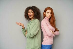 Young pretty dark haired curly woman with dark skin looking happily at camera and keeping palm raised while her unhappy redhead friend pouting sadly her lips, isolated over grey background photo