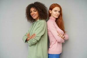 Charming young lovely ladies dressed in casual clothes folding their hands on chests and looking positively at camera with gentle smile, isolated over grey background photo